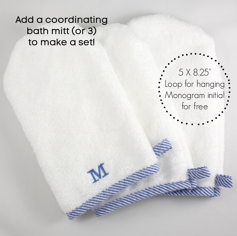 May include: Three white bath mitts with blue and white striped trim. The mitts are stacked on top of each other. The top mitt has the letter "M" embroidered on it. The text "5 X 8.25" Loop for hanging Monogram initial for free" is written in a circle around the mitts.