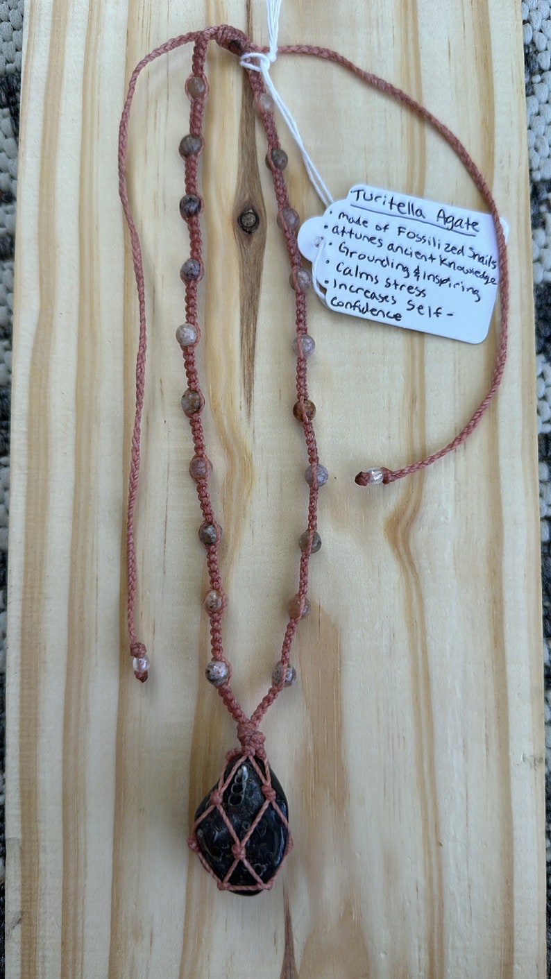 Pink necklace with neutral beads lining all the way up the neckline with 5 knots between each bead. The Agate is caged in the necklace and it is a dark black/deep green with fossilized snails that are in the front of the stone.