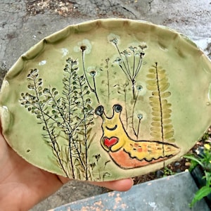 May include: An oval-shaped ceramic dish with a light green glaze, featuring an embossed illustration of a yellow slug holding a small red heart. The design includes detailed impressions of various wildflowers and fern leaves across the surface.