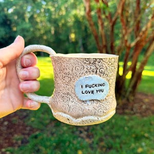 May include: A beige ceramic mug with a textured, swirling embossed pattern across the surface. A smooth, light-colored oval patch is centered on the front, featuring the black stamped text "I FUCKING LOVE YOU" in a simple, sans-serif font.