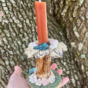 May include: A ceramic candle holder with a tree trunk design, featuring a bird perched on a flower, surrounded by pink and white flowers. The holder has a base with pink hearts and white flowers. An orange candle is placed in the holder.