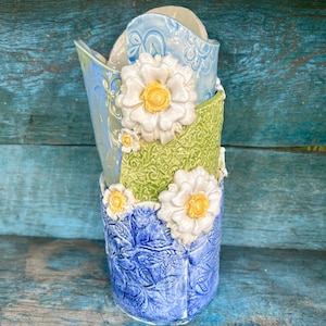 Peut inclure: Un vase en céramique artisanal à motif floral. Le vase présente un design superposé avec des couleurs bleu, vert et blanc. De grandes fleurs blanches avec des centres jaunes ornent le vase, ajoutant une touche d'élégance. Le vase a une surface texturée.