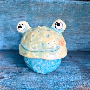 Puede incluir: Un recipiente de cerámica con forma de rana, con una cabeza verde claro y amarilla y una base azul. La rana tiene ojos grandes y expresivos y una boca sonriente. El recipiente es probablemente hecho a mano y tiene un diseño caprichoso.
