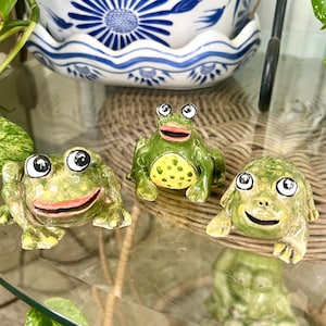 Może przedstawiać: Trzy ceramiczne figurki żab w kolorze zielonym z dużymi oczami i uśmiechniętymi pyszczkami. Żaby znajdują się na szklanej powierzchni, a w tle widać pleciony kosz i niebiesko-biały ceramiczny garnek.