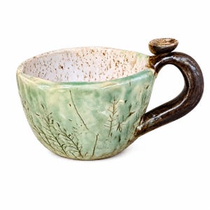 May include: A handmade ceramic mug with a light green glazed exterior featuring etched botanical patterns. The interior is white with brown speckles, and the mug has a dark brown, curved handle with a small, circular thumb rest on top.