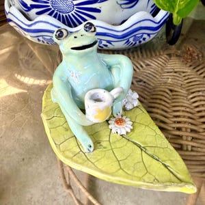 May include: A light green ceramic frog figurine sitting on a large, textured green leaf. The frog has dark eyes and a wide mouth, holding a small white mug with a yellow star design. Two small white daisy flowers with orange centers are attached to the leaf.