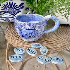 May include: A handmade blue ceramic mug with a textured surface and a handle, featuring the embossed text I GIVE A FUCK on the front. Seven small, oval-shaped blue ceramic tokens are arranged below the mug, each stamped with the word FUCK.