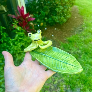 May include: A ceramic frog figurine sitting on a green leaf-shaped dish. The frog is green with black eyes and a white belly. The leaf is green with a light green vein pattern.