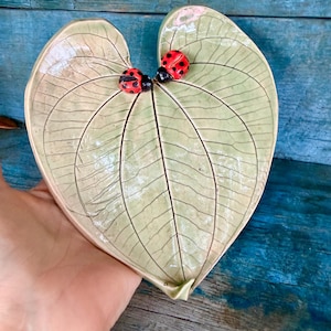 May include: A heart-shaped ceramic dish designed to resemble a light green leaf with detailed dark green veining. Two small, three-dimensional red ladybugs with black spots are attached to the upper surface of the leaf-shaped tray.