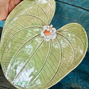 May include: A ceramic dish shaped like a large, light green lily pad with a glossy, crackled glaze finish and prominent dark vein details. A small, three-dimensional white flower with an orange center is attached to the center of the leaf.