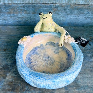 May include: A ceramic frog figurine perched on a blue and white bowl. The frog is light green with dark spots and is holding a white flower. The bowl has a textured rim and a blue and white interior.