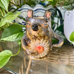 May include: A whimsical ceramic mug shaped like a bear with a sunflower design. The mug is primarily brown with pink accents on the ears and a red center on the sunflower. It has a handle and is likely used for beverages.