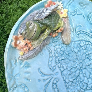 May include: A ceramic bird bath with a blue glaze and a sculpted frog with wings and flowers. The frog is sleeping with its eyes closed.