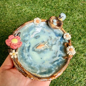 Op de afbeelding: Een handgemaakte keramische schaal met een vijverontwerp. De schaal heeft een blauwgroen interieur met een kleine oranje vis. Een vogel zit in een nest met eieren en bloemen versieren de rand. De schaal wordt tegen een groene achtergrond gehouden.