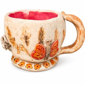 May include: A ceramic mug with a cream-colored exterior and a vibrant pink interior. The mug features raised, textured relief designs of orange butterflies, wheat stalks, and small flowers around the base. It has a thick, branch-like handle.
