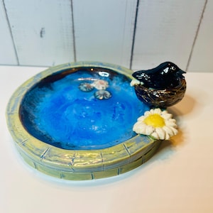 May include: A handcrafted ceramic bird bath with a vibrant blue interior, resembling a pond. A black bird sits in a nest on the side, with a white and yellow daisy nearby. The bird bath has a textured, light green and yellow rim.