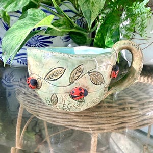 May include: A handmade ceramic mug with a light green glaze, featuring embossed leaf patterns and three small, three-dimensional red and black ladybug figures attached to the exterior. The interior of the mug is finished in a smooth, light blue glaze.