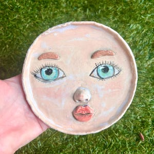 May include: A round, ceramic trinket dish with a face design. The dish features a light pink base color with hand-painted details including blue eyes, brown eyebrows, freckles, and red lips. The dish is held in a hand.