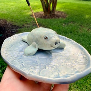May include: A light blue ceramic incense holder shaped like a manatee resting on an oval tray with a textured, mottled surface. The manatee has dark eyes and a small mouth, with a black incense stick inserted into its back, held upright.