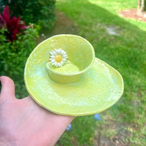 May include: A small, green ceramic cup and saucer set with a white daisy flower accent. The set has a textured, glazed finish.