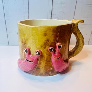 Könnte beinhalten: Eine skurrile Keramiktasse mit einer strukturierten, baumartigen Außenseite in Gelb- und Brauntönen. Zwei leuchtend rosa Schneckenfiguren mit Kulleraugen sind vorne angebracht. Der Griff hat eine geschwungene, organische Form.