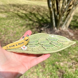 May include: A ceramic ashtray shaped like a leaf, with the word "FUCK" repeated on the leaf. A yellow slug with the words "I GIVE A FUCK" is on the ashtray. A small white flower is also on the ashtray.