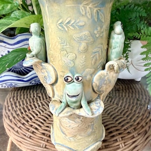 May include: A cylindrical ceramic vase with a pale yellow and blue glaze, featuring embossed patterns of mushrooms, leaves, and flowers. Three sculpted green frog figures are attached to the exterior, each resting on a leaf-shaped protrusion.