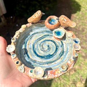 May include: Handmade ceramic bird bath with a spiral design in shades of blue and tan. Two small birds and a nest are perched on the rim. The rim is decorated with small, textured embellishments. A unique, decorative piece for the garden.