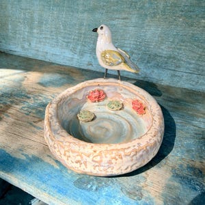 May include: A ceramic bird bath with a white and yellow bird perched on the edge. The bird bath is round with a light blue interior and decorative flowers. The bird bath is sitting on a weathered blue wooden surface.