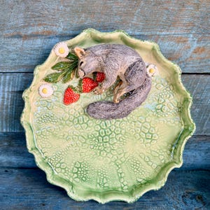 May include: A ceramic serving plate with a light green, textured surface and a scalloped edge. A gray squirrel figurine rests on the plate, next to red strawberries and white daisy accents. The plate is approximately 10 inches in diameter.