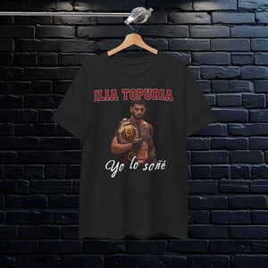 Pode incluir: Camiseta preta com uma imagem de Ilia Topuria, um lutador de artes marciais mistas, segurando um cinturão de campeonato. O texto "Ilia Topuria" e "Yo lo soñé" está impresso na camiseta.