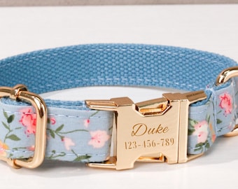 May include: A light blue dog collar with a floral pattern of pink flowers and green leaves. The collar has gold-colored metal hardware, including a buckle and a D-ring. The name "Duke" and a phone number are engraved on the buckle.