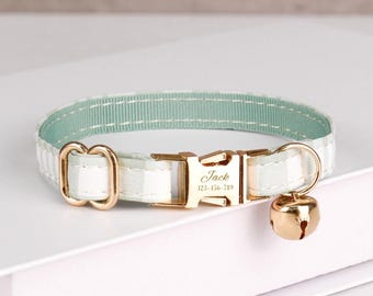 May include: A cat collar with a gold-colored buckle and bell. The collar is white with a light green stripe and a gold-colored buckle. The buckle is engraved with the name "Jack" and a phone number.