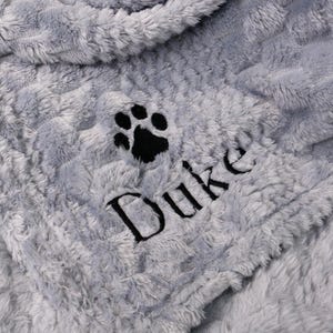 Op de afbeelding: Een zachte, grijze deken voor huisdieren met een zachte textuur. De deken heeft een zwarte pootafdruk en de naam "Duke" in zwart geborduurd. De deken is ontworpen voor comfort en warmte.