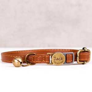 May include: A brown leather cat collar with a gold-coloured bell and buckle. The collar has a small, round tag with the name "Jack" and a phone number. The collar is designed for a pet.