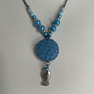 Op de afbeelding: Een blauwe ketting met een ronde hanger met een vis-schubbenpatroon en een zilveren vis-bedel. De ketting is gemaakt van blauwe en witte kralen.