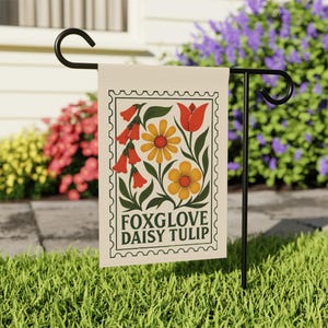 May include: A garden flag with a floral design featuring foxglove, daisy, and tulip illustrations. The flag has a cream background with a green border and the words "FOXGLOVE DAISY TULIP". The flag is displayed on a black metal stand in a garden.
