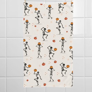 May include: A cream-colored Halloween-themed towel featuring a repeating pattern of dancing skeletons holding pumpkins. The skeletons are black and white, and the pumpkins are orange. Small orange pumpkins and stars are scattered across the background.