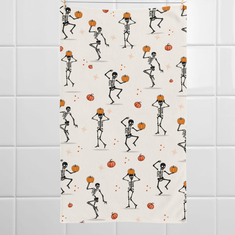 May include: A cream-colored Halloween-themed towel featuring a repeating pattern of dancing skeletons holding pumpkins. The skeletons are black and white, and the pumpkins are orange. Small orange pumpkins and stars are scattered across the background.