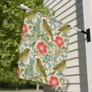May include: A decorative outdoor flag featuring a repeating pattern of green birds, red flowers, and blue foliage on a cream background. The words "Fuck Trump" are printed on the flag.