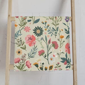 May include: A floral patterned towel with a light beige background. The towel features various flowers in shades of pink, green, and white, with the words "fuck trump" written in cursive. The towel is hanging on a wooden ladder.