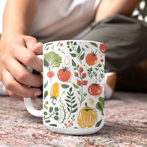 May include: White ceramic mug featuring a colorful pattern of vegetables and foliage, including tomatoes, radishes, and squash. The design incorporates the text "fuck trump" in a handwritten style. The mug has a white handle and is held by a person.