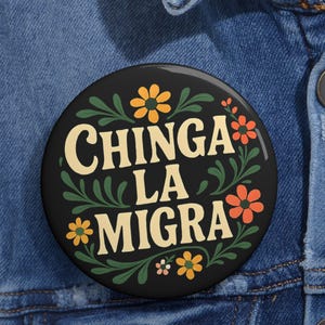 May include: A round black button with the text "CHINGA LA MIGRA" in cream-colored letters. The button is decorated with orange and yellow flowers and green leaves. The button is pinned to a blue denim jacket.