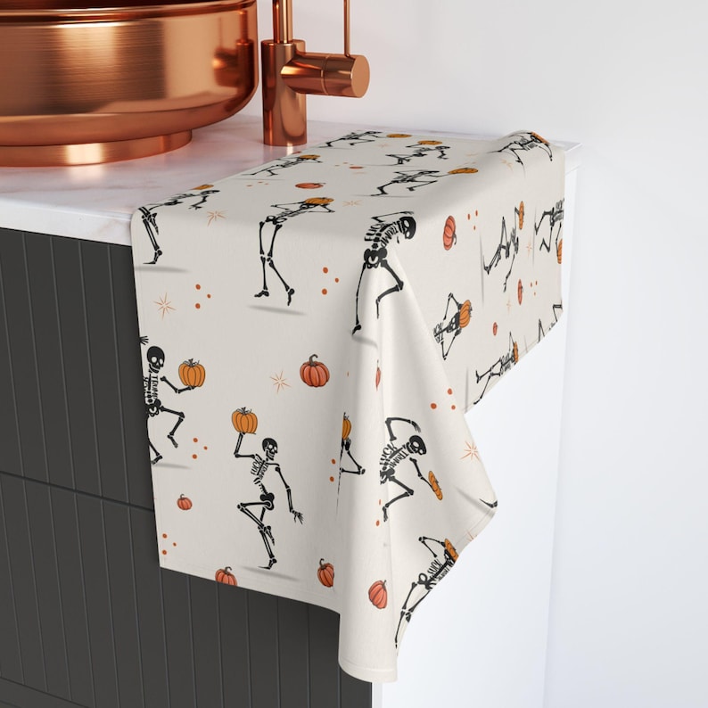 May include: A cream-colored Halloween hand towel featuring a repeating pattern of black skeletons holding pumpkins. The design includes orange pumpkins and small orange dots. The towel is draped over a countertop near a copper faucet and sink.