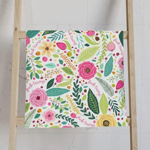 May include: A white towel with a floral pattern featuring pink roses, yellow flowers, and green leaves. The words "fuck trump" are repeated throughout the design. The towel is hanging on a wooden rack.