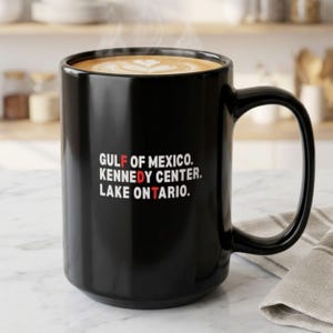 May include: A black ceramic mug filled with coffee featuring white text that reads GULF OF MEXICO. KENNEDY CENTER. LAKE ONTARIO. with the letters F, D, and T highlighted in red. The mug has a large handle and is filled with a latte with foam art.