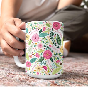 May include: White ceramic mug with a floral pattern of pink roses, green leaves, and small yellow flowers. The words "fuck trump" are interspersed throughout the design. The mug is held by a person.