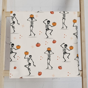 May include: A Halloween-themed towel featuring a repeating pattern of black skeletons holding orange pumpkins. The skeletons are dancing on a cream-colored background, with scattered orange pumpkins and starburst accents. The towel is displayed on a wooden rack.