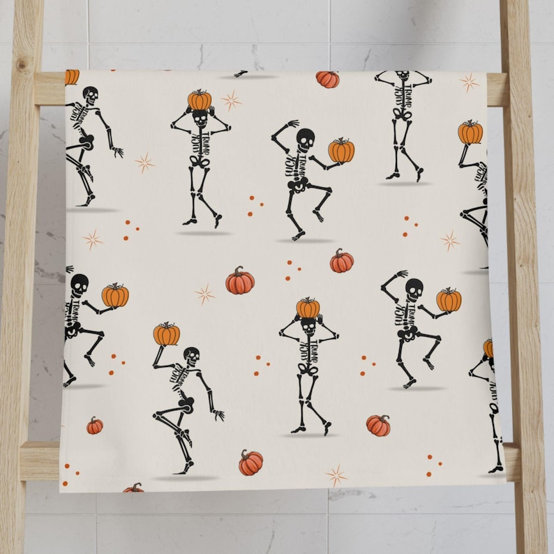 May include: A Halloween-themed towel featuring a repeating pattern of black skeletons holding orange pumpkins. The skeletons are dancing on a cream-colored background, with scattered orange pumpkins and starburst accents. The towel is displayed on a wooden rack.