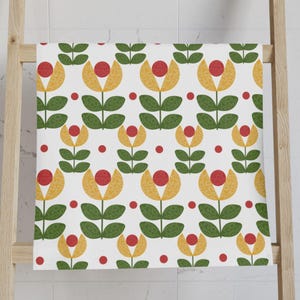 May include: A white towel with a repeating floral pattern. The design features yellow and green stylized flowers with red centers and green leaves. The towel is draped over a wooden rack.